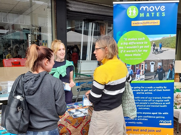 Photo of Elaine smiling and chatting at Meanwood Market with two women, Move Mates poster is next to her.