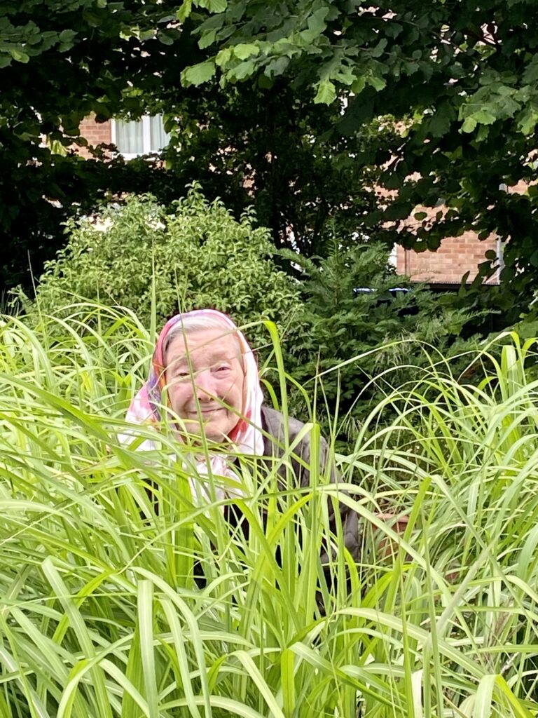 Sheila, wearing a headscarf and smiling, amongst the grasses